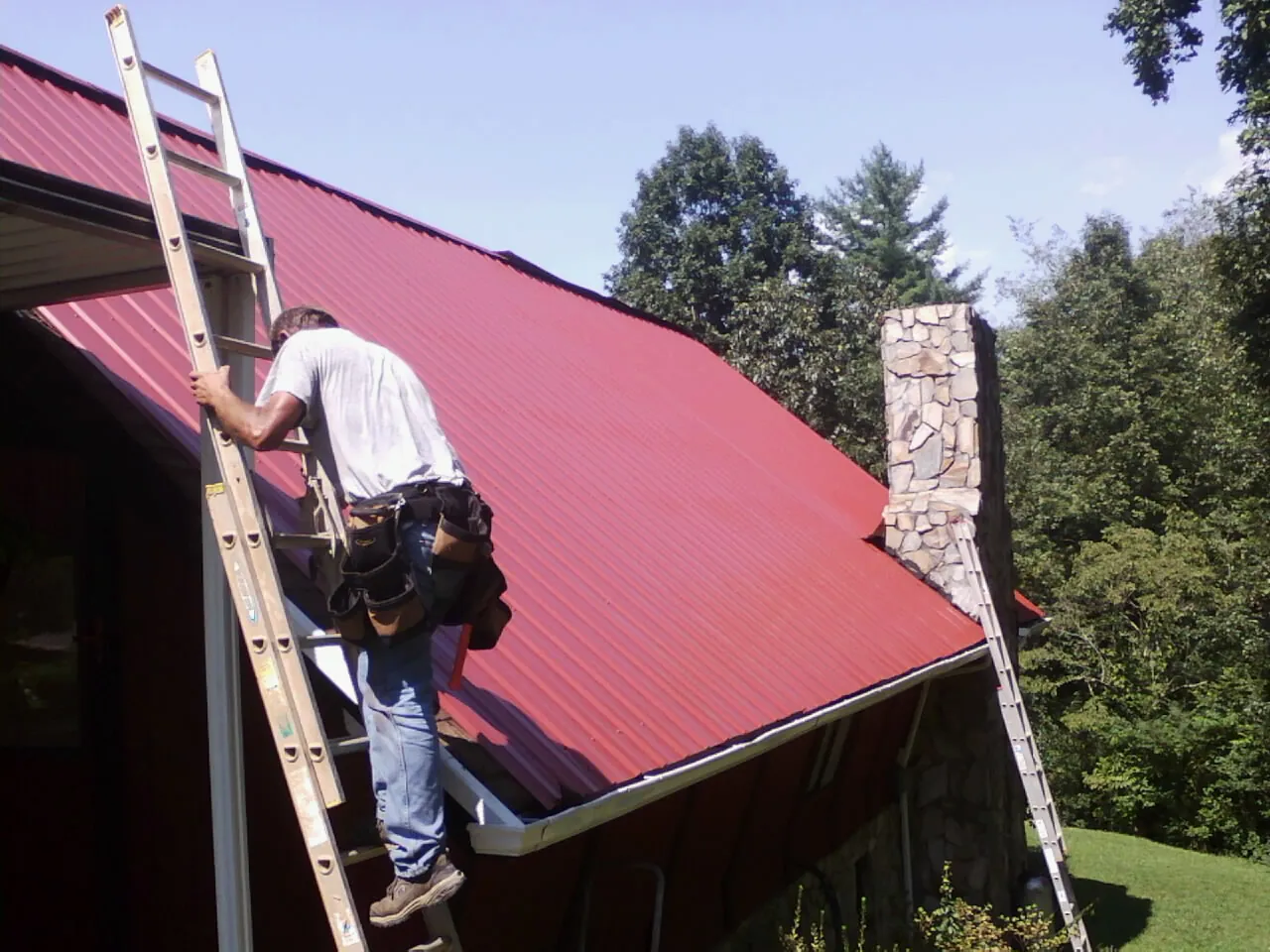 Roofer on a ladder beside a red metal roof and stone chimney — Soffit & Fascia Repair by Roofing Brothers in New Meadows